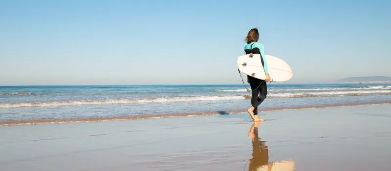 Womans wetsuit - woman walking in a wetsuit with a surfboard along the shoreline 
